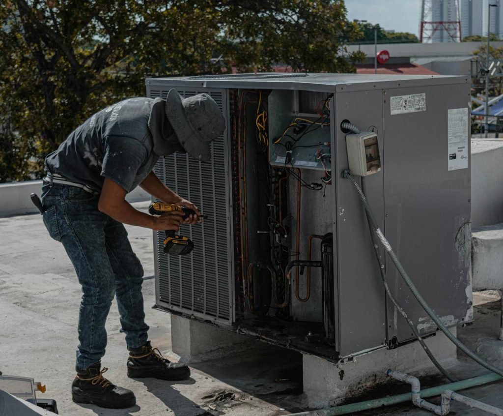 repairman-repairing-a-air-conditioner-5463587 A worker in a bucket hat repairs an outdoor air conditioning unit on a rooftop.